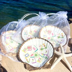 May include: Four seashell coasters with gold trim, each featuring a colorful Mahjong tile design. The coasters are packaged in sheer white drawstring bags, with a starfish and decorative wooden balls nearby.