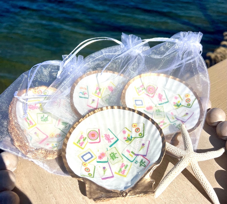 May include: Four seashell coasters with gold trim, each featuring a colorful Mahjong tile design. The coasters are packaged in sheer white drawstring bags, with a starfish and decorative wooden balls nearby.