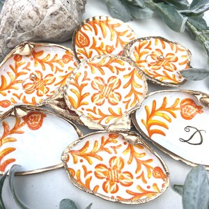 Könnte beinhalten: Handbemalte Austernschalen-Ringschalen mit goldenem Rand. Die Schalen haben einen weißen Hintergrund mit orangefarbenen floralen Designs. Eine Schale hält einen Ring. Die Schalen sind auf einer weißen Oberfläche mit Grün angeordnet.