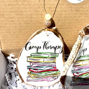 May include: A white oyster shell ornament with gold accents, featuring a stack of colorful books and the text "Group Therapy".