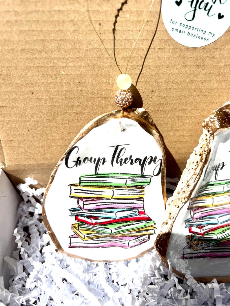 May include: A white oyster shell ornament with gold accents, featuring a stack of colorful books and the text "Group Therapy".