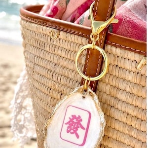 May include: A gold-colored keychain with a white, irregular-shaped charm. The charm has a pink and white design with a Chinese character. The keychain is attached to a brown leather strap on a woven straw bag.