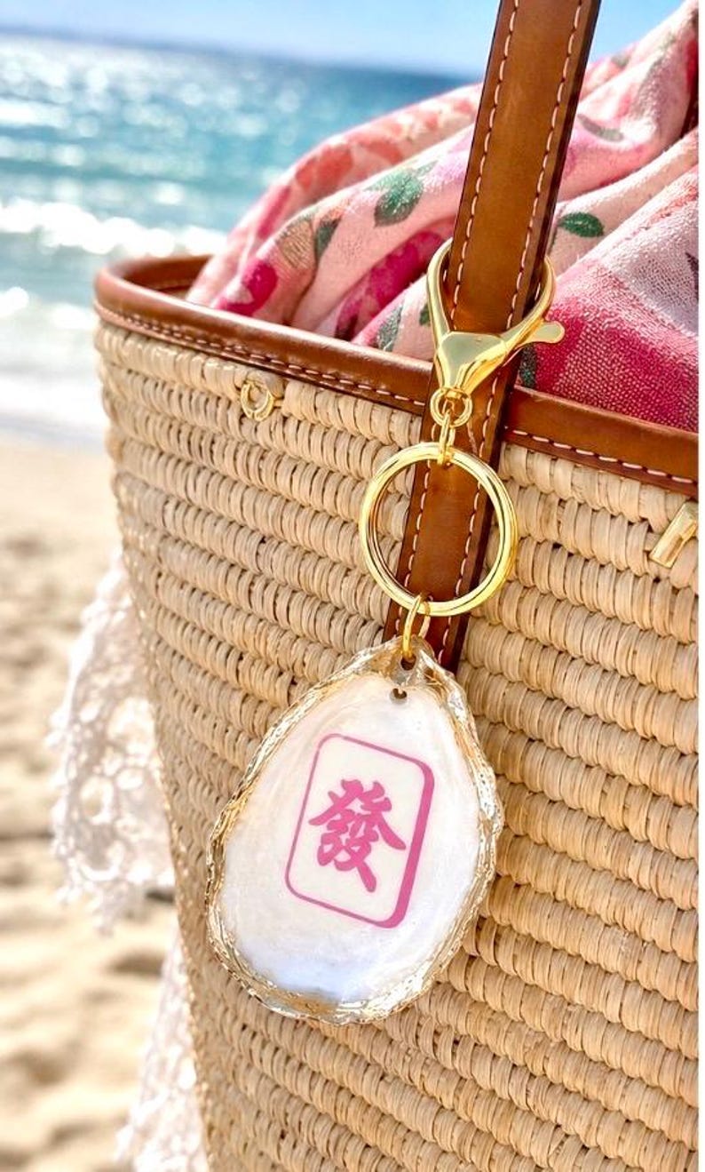 May include: A gold-colored keychain with a white, irregular-shaped charm. The charm has a pink and white design with a Chinese character. The keychain is attached to a brown leather strap on a woven straw bag.