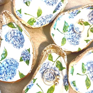 May include: Oyster shell ring dishes with gold edging and a blue and white hydrangea floral pattern. Some dishes hold rings. The shells are arranged on a light brown surface.
