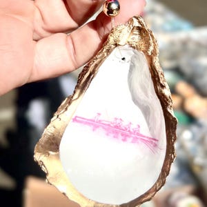 May include: An oyster shell pendant with a gold-painted edge and a pink rowing team design inside. The pendant is strung on a gold-colored cord with a pink and gold bead accent.