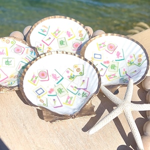 May include: Four white scallop shell coasters with gold rims, each decorated with colorful Mahjong tile designs. A white starfish and wooden beads are also visible, suggesting a beach or coastal theme.