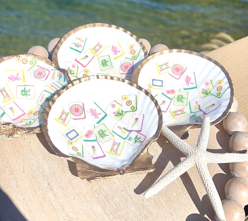 May include: Four white scallop shell coasters with gold rims, each decorated with colorful Mahjong tile designs. A white starfish and wooden beads are also visible, suggesting a beach or coastal theme.