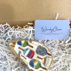 May include: A white oyster shell dish with a gold rim, featuring a colorful pattern of paddleball rackets. The dish is sitting on a bed of white shredded paper.