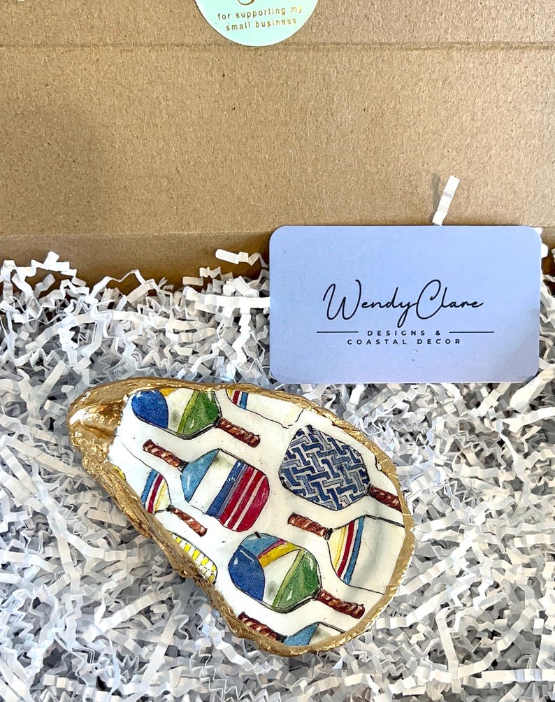 May include: A white oyster shell dish with a gold rim, featuring a colorful pattern of paddleball rackets. The dish is sitting on a bed of white shredded paper.