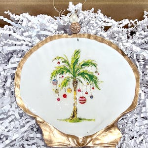 May include: A white seashell ornament with a gold rim, featuring a painted palm tree decorated with colorful Christmas ornaments. The ornament has a pearl and gold string for hanging.