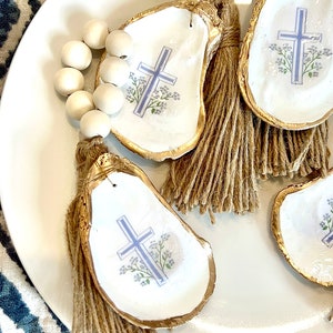 May include: Four white oyster shells with gold trim and a blue cross and floral design. Each shell has a brown tassel attached. A string of white beads is in the background.
