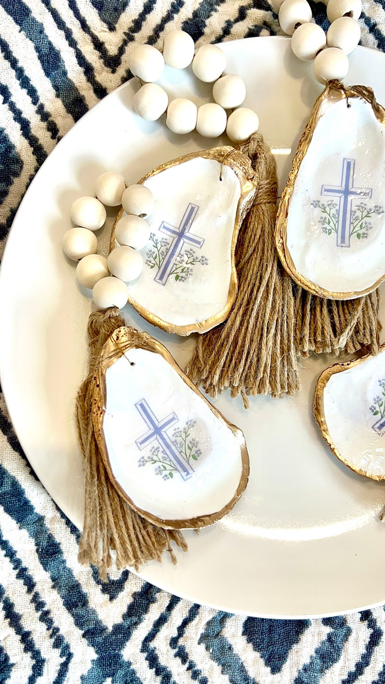 May include: Four white oyster shells with gold trim and a blue cross and floral design. Each shell has a brown tassel attached. A string of white beads is in the background.