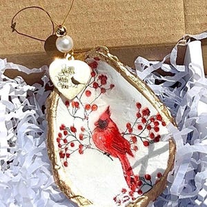 May include: Oyster shell ornament featuring a red cardinal bird perched on a branch with red berries. The shell has a gold-painted edge and a heart-shaped charm that reads "I am Always With You". A pearl and gold wire complete the design.