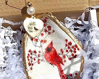 May include: Oyster shell ornament featuring a red cardinal bird perched on a branch with red berries. The shell has a gold-painted edge and a heart-shaped charm that reads "I am Always With You". A pearl and gold wire complete the design.