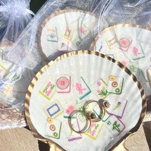 May include: A white seashell ring dish with a gold-painted rim, decorated with colorful Mahjong tile designs. Two gold rings with gemstones rest inside the dish. Several similar dishes are visible, some in sheer white bags.