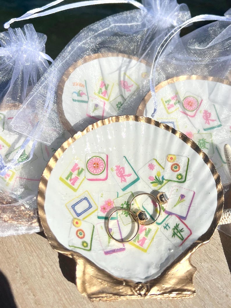 May include: A white seashell ring dish with a gold-painted rim, decorated with colorful Mahjong tile designs. Two gold rings with gemstones rest inside the dish. Several similar dishes are visible, some in sheer white bags.
