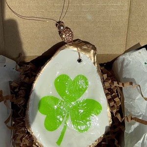 May include: A white oyster shell ornament with a gold rim and a green shamrock painted on the front. The ornament is hanging from a gold wire with a gold bead.