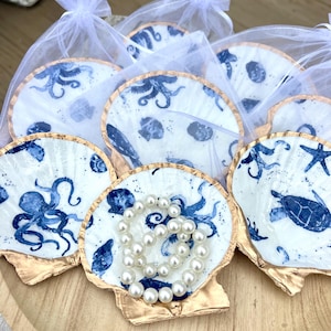 May include: A collection of white scallop shells decorated with blue marine life illustrations, including octopuses, sea turtles, and starfish. Each shell features a metallic gold painted edge and is individually wrapped in a white organza bag.