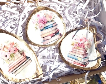 Peut inclure: Trois ornements en coquille d'huître avec des bords dorés, chacun représentant une pile de livres et des arrangements floraux roses. Les ornements sont blancs avec une bordure dorée, présentés dans une boîte avec du papier de remplissage blanc.