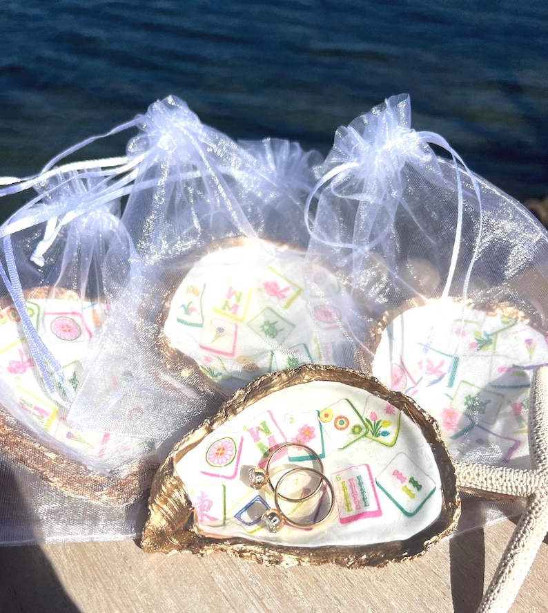 Pu&ograve; includere: Piatti portanelli a conchiglia con bordi dorati, ognuno con un anello. I piatti sono decorati con un motivo colorato e sono presentati in piccoli sacchetti bianchi trasparenti. Lo sfondo mostra uno specchio d'acqua.