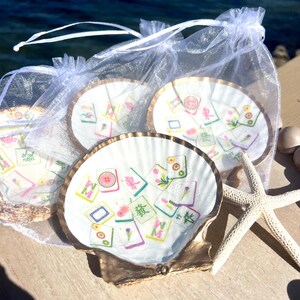 May include: A set of four seashell coasters with a gold trim, each featuring a colorful Mahjong tile design. The coasters are presented in a sheer white drawstring bag, with a starfish nearby. The coasters are on a wooden surface.
