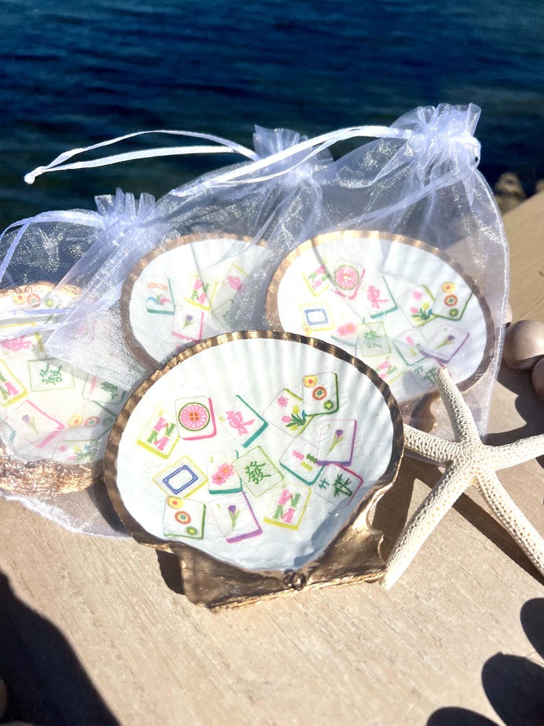May include: A set of four seashell coasters with a gold trim, each featuring a colorful Mahjong tile design. The coasters are presented in a sheer white drawstring bag, with a starfish nearby. The coasters are on a wooden surface.