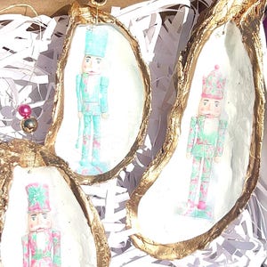 May include: Three oyster shell ornaments with gold-painted edges and a white interior. Each shell features a miniature nutcracker soldier in shades of blue, pink, and green. The ornaments are suspended by a string with a bead.