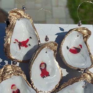 Könnte beinhalten: Fünf goldbemalte Austernmuschel-Ornamente mit weißen Innenräumen. Jedes Ornament zeigt ein handgemaltes Bild vom Weihnachtsmann in einer anderen Pose. Die Ornamente sind mit goldfarbenem Draht aufgefädelt und haben eine silberne Perle am Ende.