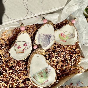 May include: Four oyster shell ornaments with gold trim and painted teacups. Each ornament has a pink bead and a gold loop for hanging.