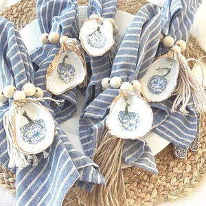 May include: Four blue and white striped napkins with a white oyster shell with a blue and white floral design and a brown tassel. The oyster shells are tied to the napkins with wooden beads.