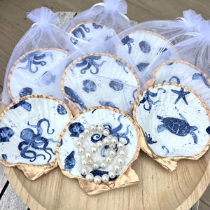 May include: A collection of seashell-shaped trinket dishes with blue and white sea creature designs, including octopuses and turtles. Each dish has a gold-painted edge and is presented in a sheer white bag. A pearl necklace rests on one dish.