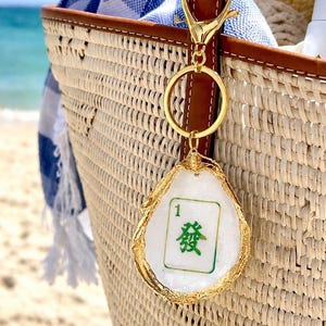 May include: A gold-colored keychain with a white and green Mahjong tile charm. The charm is in an oyster shell shape with gold trim. The keychain is attached to a woven straw tote bag with a blue and white striped towel inside.
