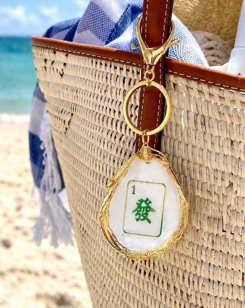 May include: A gold-colored keychain with a white and green Mahjong tile charm. The charm is in an oyster shell shape with gold trim. The keychain is attached to a woven straw tote bag with a blue and white striped towel inside.