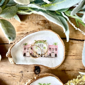 May include: Two gold-rimmed oyster shell trinket dishes with a white background and a painted design of pink buildings and palm trees. One dish has a pair of gold hoop earrings inside.