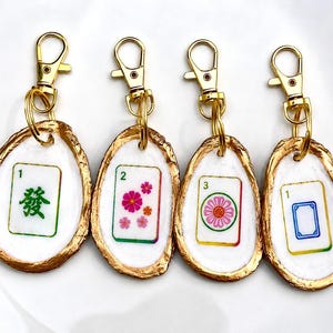 May include: Four keychains featuring Mahjong tile designs. Each tile is set in a white oyster shell with a gold-painted edge and a gold-colored clasp. The tiles display various symbols and numbers in green, pink, and blue.