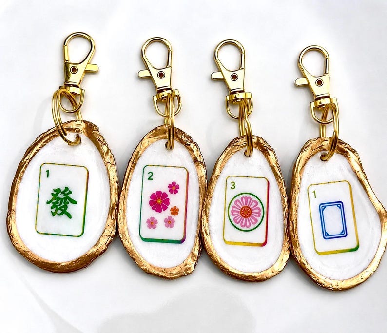 May include: Four keychains featuring Mahjong tile designs. Each tile is set in a white oyster shell with a gold-painted edge and a gold-colored clasp. The tiles display various symbols and numbers in green, pink, and blue.