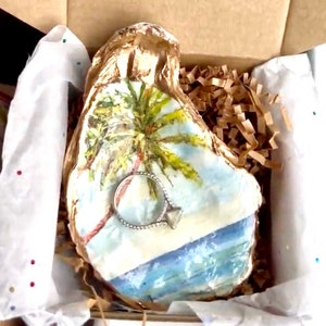 May include: A white oyster shell with a painted scene of a palm tree and ocean waves. A silver ring with a diamond is resting on the shell.