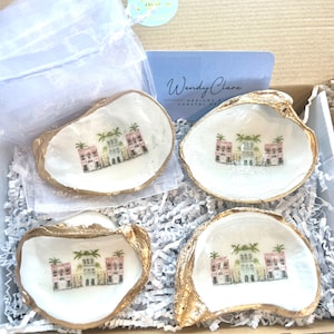 May include: Set of four white oyster shell dishes with gold trim. Each dish features a hand-painted design of a colorful building with palm trees. The dishes are packaged in a box with shredded paper.