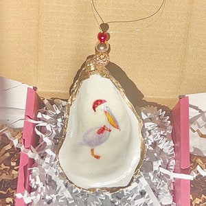 May include: A white oyster shell ornament with a gold rim and a painted pelican wearing a red Santa hat. The ornament is hanging from a gold wire with red beads.