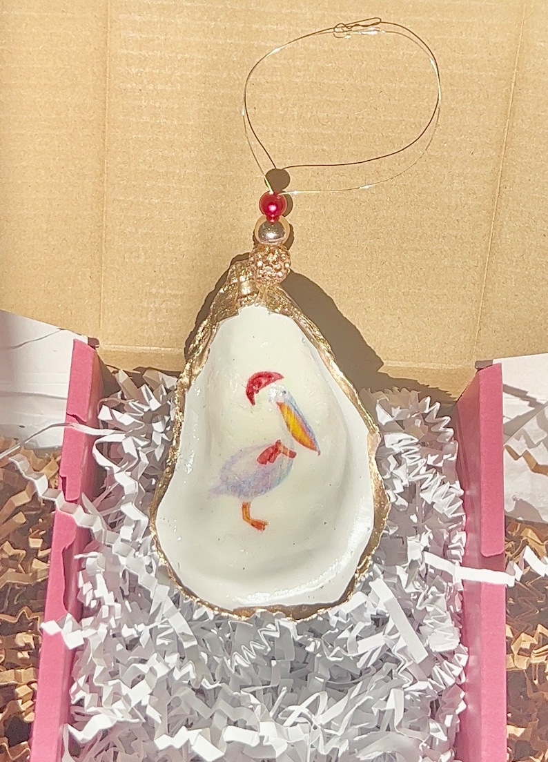 May include: A white oyster shell ornament with a gold rim and a painted pelican wearing a red Santa hat. The ornament is hanging from a gold wire with red beads.