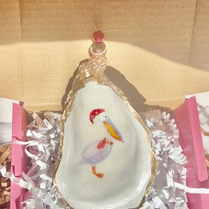 May include: A white oyster shell ornament with a gold rim, featuring a hand-painted pelican with a red Santa hat and yellow beak. The ornament is hanging from a gold string with red and gold beads.