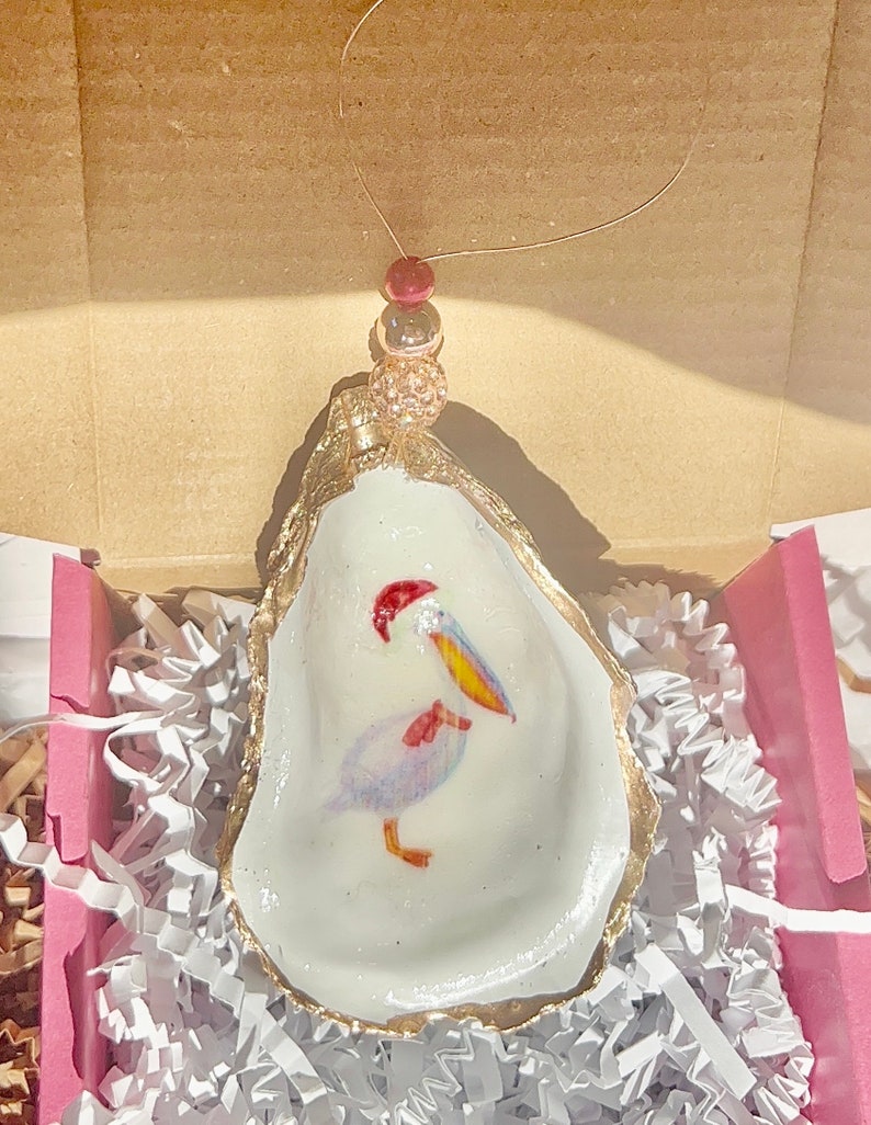 May include: A white oyster shell ornament with a gold rim, featuring a hand-painted pelican with a red Santa hat and yellow beak. The ornament is hanging from a gold string with red and gold beads.