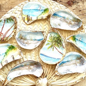 May include: A collection of eight oyster shells painted with various beach scenes. The shells are arranged in a circle on a woven straw background. The scenes include palm trees, seashells, sailboats, and beach scenes.