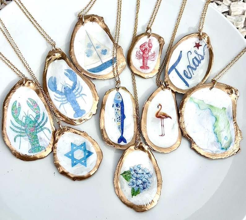 K&ouml;nnte beinhalten: Eine Sammlung von zehn Halsketten mit Austernschalen, deren R&auml;nder gold bemalt sind. Die Schalen sind mit verschiedenen handgemalten Motiven verziert, darunter ein Hummer, ein Segelboot, ein Fisch, ein Flamingo, eine blaue Hortensie, ein Davidstern und das Wort Texas.
