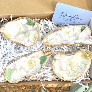 May include: Four white oyster shells with gold trim and floral designs. The shells are arranged in a box with white tissue paper. The box has a business card that reads "Wendy Clare Designs & Coastal Decor".
