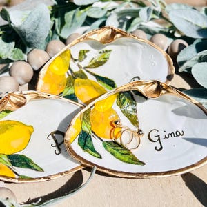 May include: Three white seashell ring dishes with gold rims, hand-painted with yellow lemons and green leaves. One dish has the name "Gina" written in black script, and another has the letter "P". Two gold rings rest in one dish.