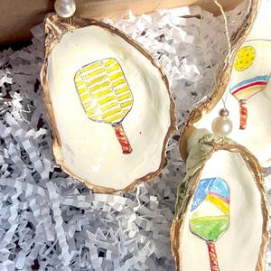May include: Three oyster shell ornaments with gold trim, each featuring a different hand-painted design of a paddleball racket. The rackets are painted in bright colors, including yellow, red, blue, and green.