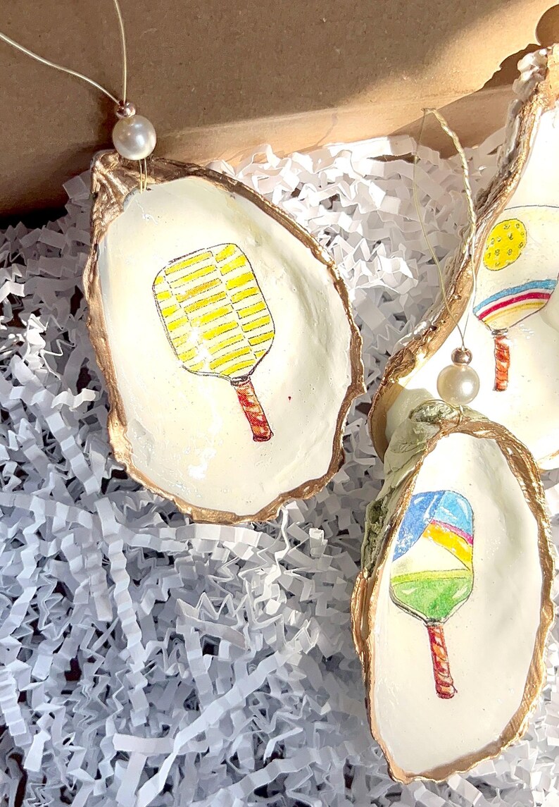 May include: Three oyster shell ornaments with gold trim, each featuring a different hand-painted design of a paddleball racket. The rackets are painted in bright colors, including yellow, red, blue, and green.
