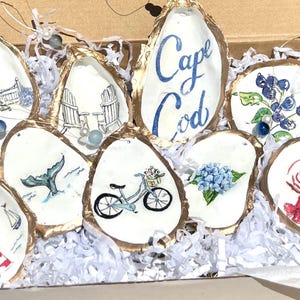 Puede incluir: Una colección de adornos de concha de ostra pintados a mano con bordes dorados, con temas costeros como un faro, una cola de ballena, una bicicleta y las palabras "Cape Cod". Cada concha es única y decorada con diseños de acuarela.