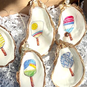 May include: Five oyster shell ornaments with gold trim, each featuring a different hand-painted design of a paddleball racket. The rackets are painted in various colors and patterns, including yellow, blue, red, and green.